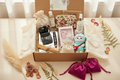 The Deep Dive — open kraft gift box with cream tissue, Chanel compact mirror and blush on etched glass tray, Love Shack Fancy makeup bag, Mofusand plush keychain, Love Shack Fancy mini perfumes, Chanel coffee table book, Voluspa candle, palo santo, lavender sachet, artisan lotion with no branding, plain muslin bag, and magenta satin drawstring bags outside